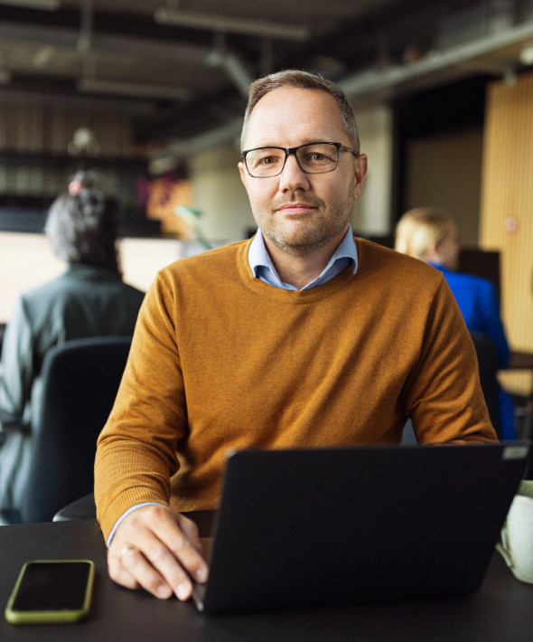 En mand sidder ved sin computer og smiler til kameraet